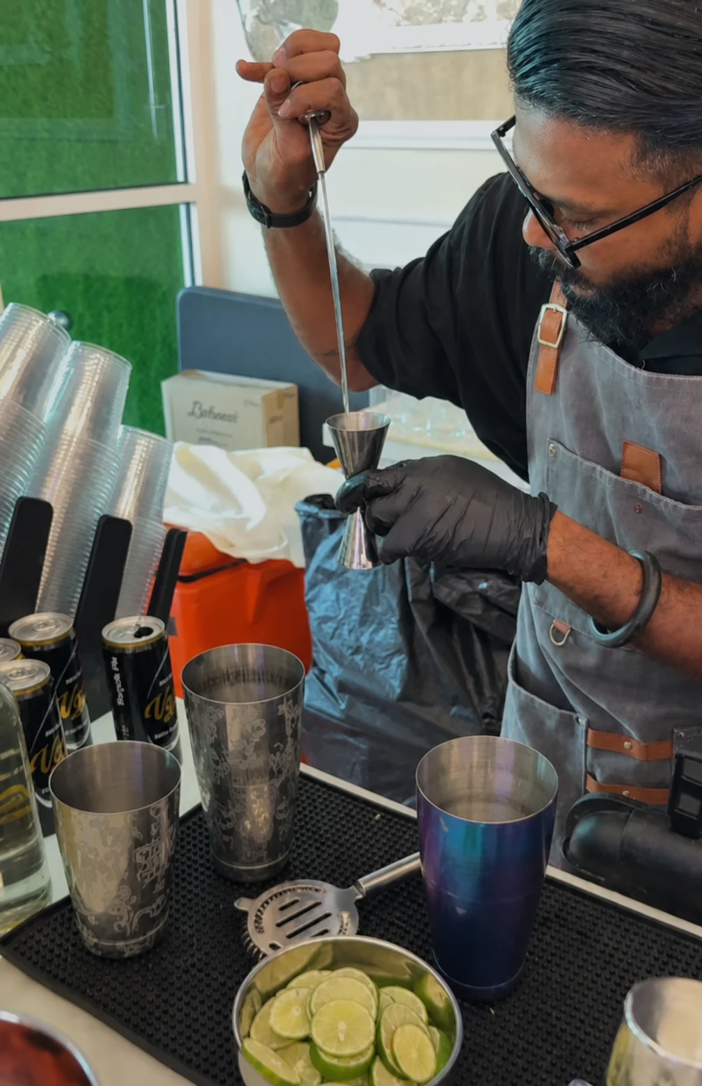 A bartender measuring a drink with a jigger beside shakers and fresh limes.